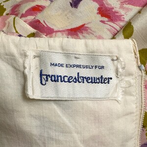 May include: Close-up of a vintage garment label. The label is white with the text "MADE EXPRESSLY FOR" above the name "francesbrewster" in dark blue script. The fabric has a floral pattern in pink, purple, and green.