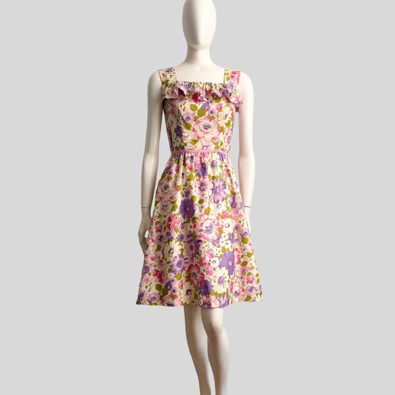 May include: A vintage floral print dress with a square neckline and a ruffled trim. The dress features a pink, purple, and green floral pattern on a cream background. It has a fitted bodice and a flared skirt, with a pink belt at the waist.