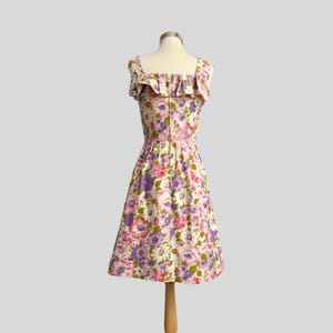 May include: A vintage floral print dress with a square neckline and ruffled trim. The dress features a pink belt and a knee-length skirt. The fabric is patterned with pink, purple, and white flowers on a cream background.