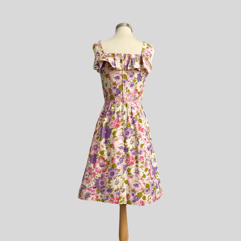 May include: A vintage floral print dress with a square neckline and ruffled trim. The dress features a pink belt and a knee-length skirt. The fabric is patterned with pink, purple, and white flowers on a cream background.