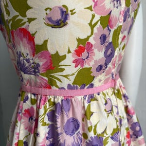 May include: A vintage floral dress featuring a vibrant pattern of large white, pink, and purple flowers with green leaves. The dress has a fitted bodice, a flared skirt, and a thin pink ribbon belt. The fabric appears to be a lightweight cotton.