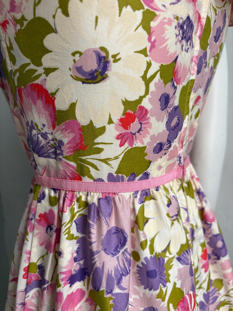 May include: A vintage floral dress featuring a vibrant pattern of large white, pink, and purple flowers with green leaves. The dress has a fitted bodice, a flared skirt, and a thin pink ribbon belt. The fabric appears to be a lightweight cotton.