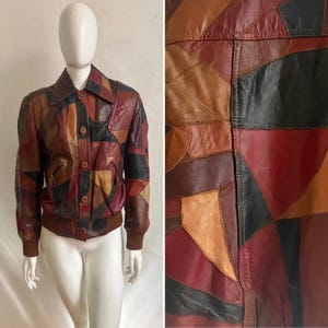 May include: A multi-colored leather jacket featuring a patchwork design of brown, red, black, and tan geometric leather pieces. The jacket includes a pointed collar, a front button closure, and brown ribbed knit cuffs and a matching ribbed hemline.