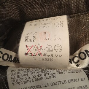 K&ouml;nnte beinhalten: Ein schwarz-wei&szlig;es Etikett mit dem Text "Commes des Garcons" und Pflegehinweisen f&uuml;r Pailletten und Perlen. Das Etikett enth&auml;lt auch den Text "D-TK 9210" und "(株)コムデギャルソン".