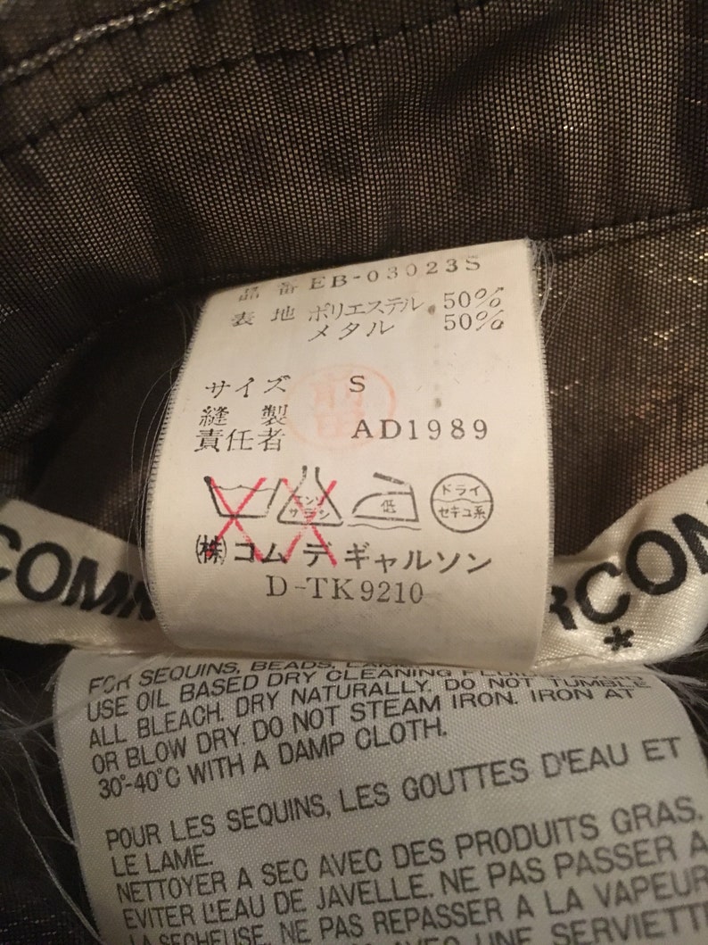 K&ouml;nnte beinhalten: Ein schwarz-wei&szlig;es Etikett mit dem Text "Commes des Garcons" und Pflegehinweisen f&uuml;r Pailletten und Perlen. Das Etikett enth&auml;lt auch den Text "D-TK 9210" und "(株)コムデギャルソン".