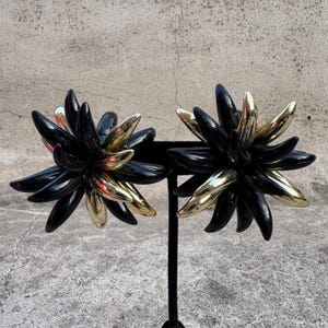 Puede incluir: Un par de pendientes negros y dorados con un diseño floral. Los pendientes están hechos de plástico negro con detalles dorados.