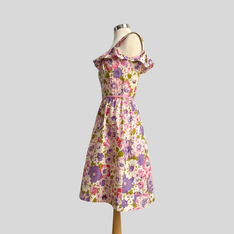 May include: A vintage floral dress with a pink belt and off-the-shoulder ruffle detail. The dress features a cream background with large pink and purple flowers and green leaves. The dress is displayed on a mannequin.