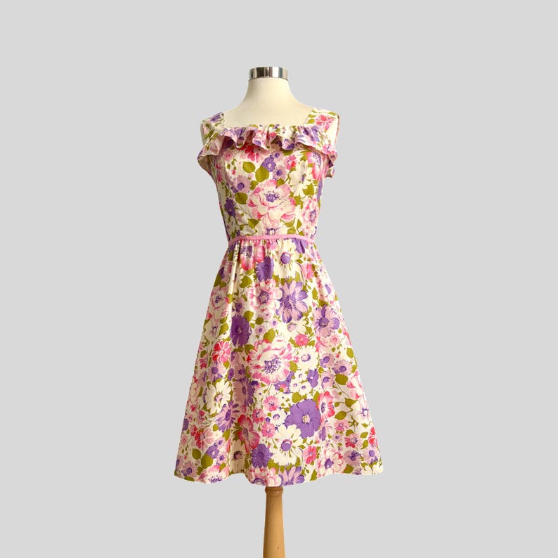 May include: A vintage floral print dress on a mannequin. The dress features a square neckline with a ruffled trim, a fitted bodice, and a flared skirt. The fabric is patterned with pink, purple, and white flowers, and a pink belt. The dress is knee-length.