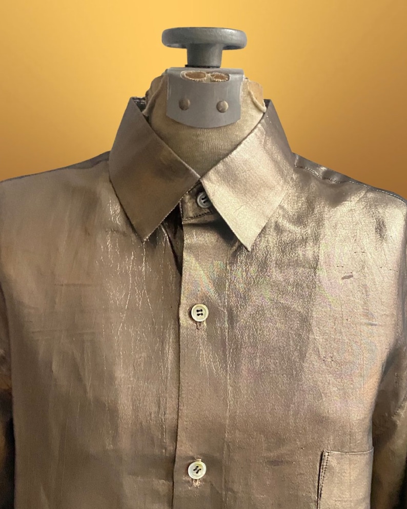 K&ouml;nnte beinhalten: Ein lang&auml;rmliges Hemd in Metallic-Gold mit Knopfleiste und einer einzigen Tasche auf der linken Seite.