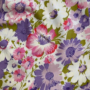 May include: Close-up of a floral fabric featuring large, stylized flowers in shades of pink, purple, and white against a white background. The flowers have green leaves and stems. The fabric has a vintage, retro aesthetic.