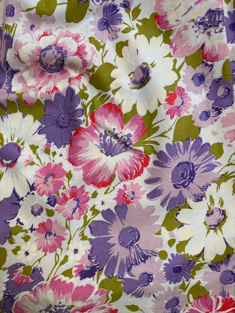 May include: Close-up of a floral fabric featuring large, stylized flowers in shades of pink, purple, and white against a white background. The flowers have green leaves and stems. The fabric has a vintage, retro aesthetic.