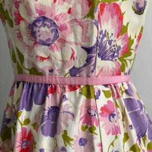 May include: A floral print dress featuring large flowers in shades of pink, purple, and white, accented with green leaves. The dress has a fitted bodice and a flared skirt, cinched at the waist with a pink ribbon. The fabric has a slightly textured appearance.