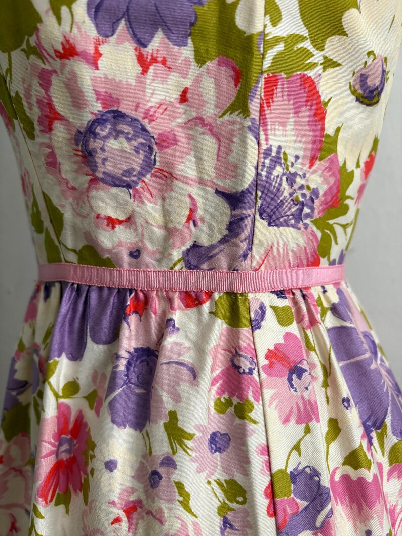May include: A floral print dress featuring large flowers in shades of pink, purple, and white, accented with green leaves. The dress has a fitted bodice and a flared skirt, cinched at the waist with a pink ribbon. The fabric has a slightly textured appearance.