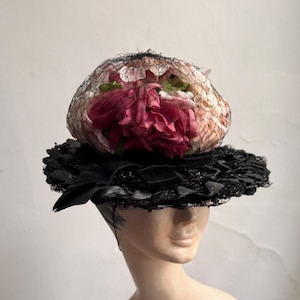 Puede incluir: Un sombrero negro y blanco con una gran flor rosa y blanca en la parte superior. El sombrero está hecho de paja y tiene un ala ancha.