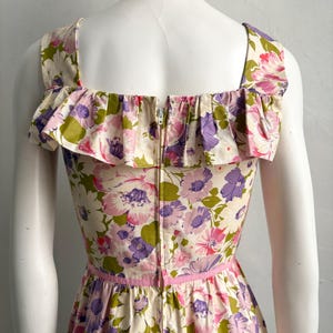 May include: A vintage floral print dress featuring a square neckline and ruffled trim. The dress showcases a vibrant pattern of pink, purple, and green flowers on an off-white background. A pink ribbon accents the waist, and a zipper runs down the back.