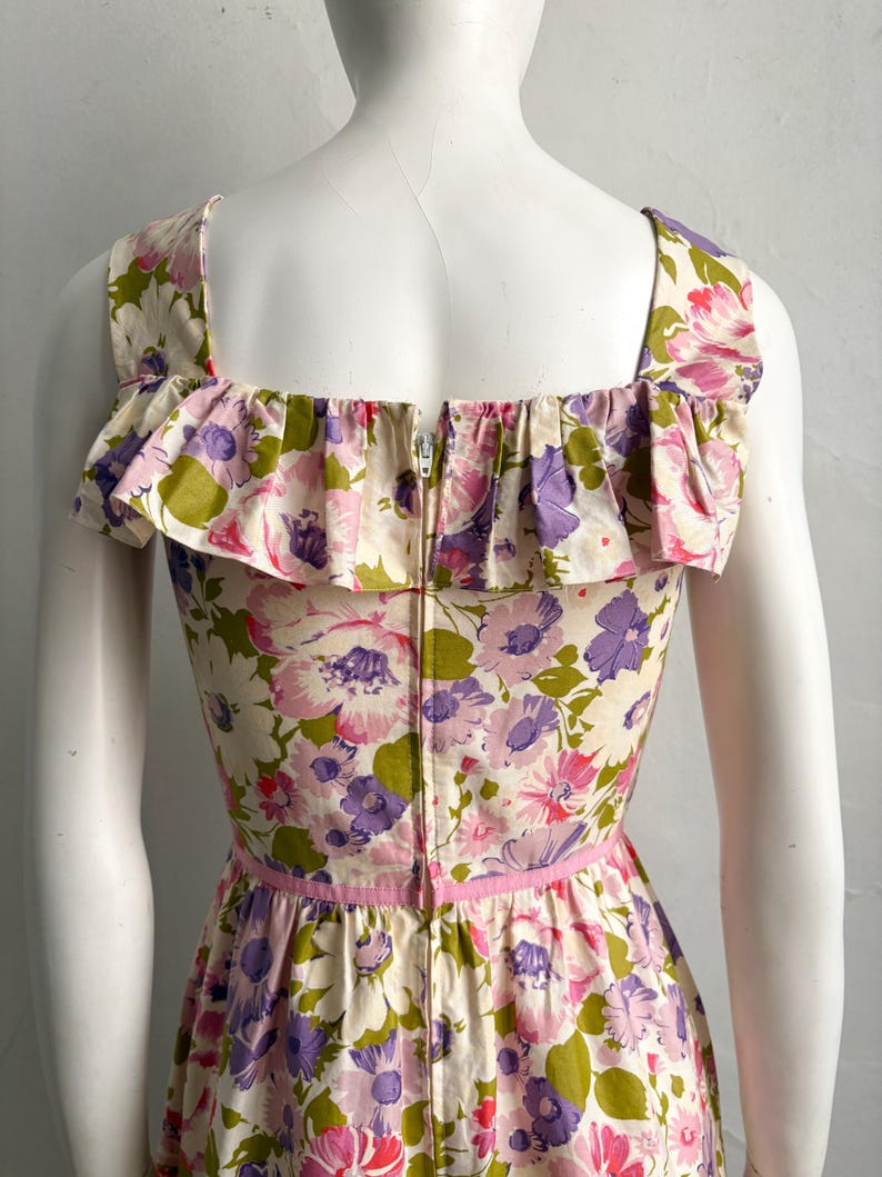 May include: A vintage floral print dress featuring a square neckline and ruffled trim. The dress showcases a vibrant pattern of pink, purple, and green flowers on an off-white background. A pink ribbon accents the waist, and a zipper runs down the back.