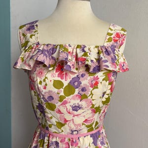 May include: A vintage floral print dress with a square neckline and ruffled trim. The dress features a pink ribbon belt and a vibrant pattern of pink, purple, and green flowers on a white background. The dress is displayed on a mannequin.
