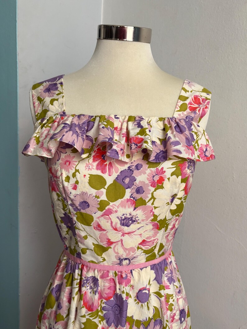 May include: A vintage floral print dress with a square neckline and ruffled trim. The dress features a pink ribbon belt and a vibrant pattern of pink, purple, and green flowers on a white background. The dress is displayed on a mannequin.