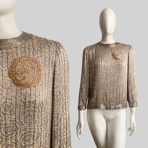 Op de afbeelding: Een vintage top met lange mouwen in lichtgrijs en goud. De top heeft een ronde halslijn en is versierd met pailletten en kralen. Een cirkelvormig kralenontwerp staat op de borst.