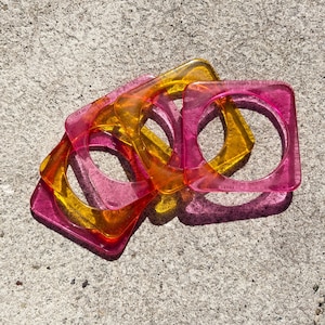 Op de afbeelding: Een verzameling doorschijnende vierkante armbanden in roze, oranje en geel. De armbanden hebben een vierkante buitenkant met een cirkelvormige opening in het midden. De sieraden liggen op een getextureerd, grijs oppervlak.