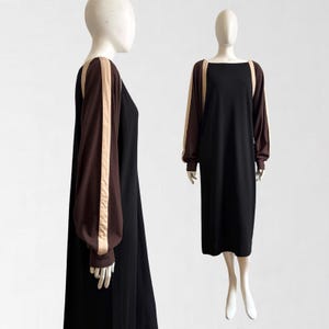 Könnte beinhalten: Ein schwarzes Midikleid mit langen, dunkelbraunen Ärmeln, die in der Mitte mit einem vertikalen beigen Streifen versehen sind. Das Kleid hat einen geraden, weiten Ausschnitt und eine lockere, fließende Passform, die unter das Knie reicht.