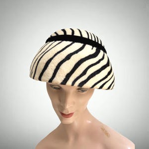 May include: A dome-shaped hat featuring a cream-colored base with black zebra-like stripes. The hat is accented with a thin, horizontal black velvet band that encircles the crown, providing a distinct contrast against the textured, patterned fabric.