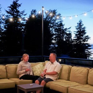May include: A couple sits on a patio with string lights overhead. They are relaxing on a light green sectional sofa. The man is wearing a white button-down shirt and the woman is wearing a gray sweater.