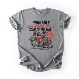 May include: Gray t-shirt with the text "PROBABLY JUST GONNA JINGLE SOME OF THE WAY I'M TIRED". The graphic shows a raccoon dressed as Santa Claus in a sleigh. The text "I'M TIRED" is in a red circle.