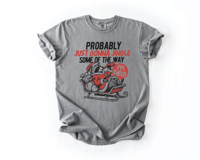 May include: Gray t-shirt with the text "PROBABLY JUST GONNA JINGLE SOME OF THE WAY I'M TIRED". The graphic shows a raccoon dressed as Santa Claus in a sleigh. The text "I'M TIRED" is in a red circle.