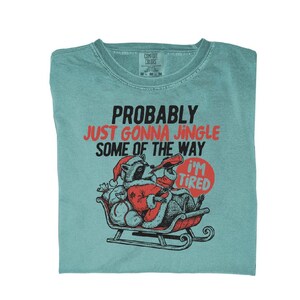 May include: A teal Comfort Colors t-shirt with the text "PROBABLY JUST GONNA JINGLE SOME OF THE WAY" in black and red. A cartoon raccoon in a Santa suit is in a sleigh, with a sign that says "I'M TIRED".