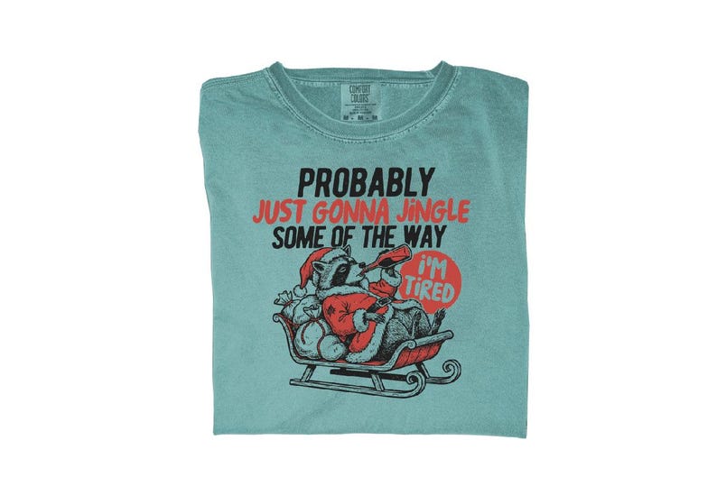 May include: A teal Comfort Colors t-shirt with the text "PROBABLY JUST GONNA JINGLE SOME OF THE WAY" in black and red. A cartoon raccoon in a Santa suit is in a sleigh, with a sign that says "I'M TIRED".