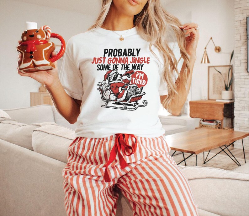 May include: White t-shirt with the text "Probably Just Gonna Jingle Some of the Way I'm Tired" and a Santa Claus graphic. The person is wearing red and white striped pajama pants and holding a gingerbread man mug.