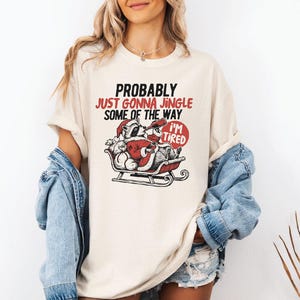 May include: Cream-colored t-shirt with a graphic of a raccoon dressed as Santa Claus in a sleigh, with the text "PROBABLY JUST GONNA JINGLE SOME OF THE WAY I'M TIRED". The design is in red and black.