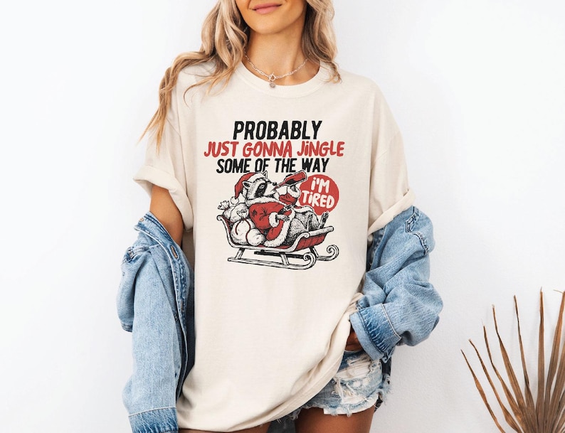 May include: Cream-colored t-shirt with a graphic of a raccoon dressed as Santa Claus in a sleigh, with the text "PROBABLY JUST GONNA JINGLE SOME OF THE WAY I'M TIRED". The design is in red and black.