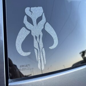 May include: A white vinyl sticker of the Mandalorian symbol, also known as the "Death Watch" symbol, on a car window. The sticker is slightly faded and has a textured surface. The text "FRG876 1-800-551-23" is visible in the lower right corner of the image.