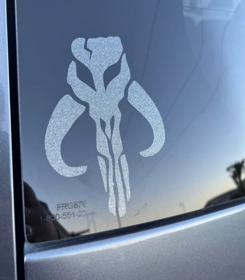 May include: A white vinyl sticker of the Mandalorian symbol, also known as the "Death Watch" symbol, on a car window. The sticker is slightly faded and has a textured surface. The text "FRG876 1-800-551-23" is visible in the lower right corner of the image.