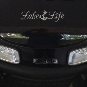May include: A black golf cart front hood featuring a white decal with the text Lake Life and an anchor symbol between the words. The vehicle has two clear headlights and a central black emblem with the silver text EZ-GO visible below the decal.