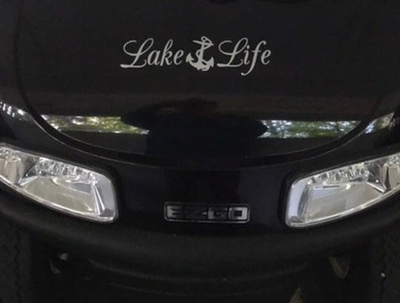May include: A black golf cart front hood featuring a white decal with the text Lake Life and an anchor symbol between the words. The vehicle has two clear headlights and a central black emblem with the silver text EZ-GO visible below the decal.