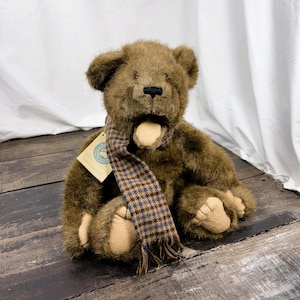 May include: A brown teddy bear with a plaid scarf. The bear has a tan muzzle and paws, and a black nose. The scarf is brown, blue, and tan. The bear is sitting on a wooden surface.