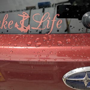May include: A rose gold vinyl decal applied to a vehicle rear window, featuring the text Lake Life in a cursive script font with an anchor icon replacing the letter k. The decal is positioned above the trunk lid of a maroon car with water droplets.