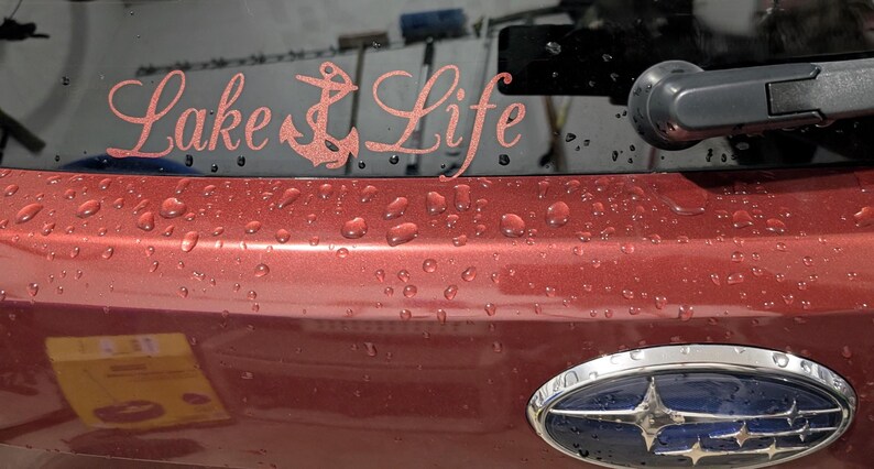 May include: A rose gold vinyl decal applied to a vehicle rear window, featuring the text Lake Life in a cursive script font with an anchor icon replacing the letter k. The decal is positioned above the trunk lid of a maroon car with water droplets.