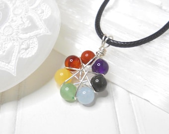 May include: A silver wire-wrapped pendant necklace featuring seven colorful gemstone beads. The beads are arranged in a circular pattern, each representing a chakra. The necklace hangs from a black cord.