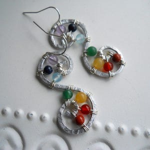 May include: Silver earrings with a swirl design and colorful gemstones. The gemstones are in a rainbow pattern, with red, orange, yellow, green, blue, indigo, and violet.