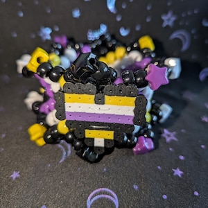 May include: A beaded bracelet featuring the non-binary pride flag colors: yellow, white, purple, and black. The bracelet is made of small, colorful beads and includes star-shaped beads. The background is a dark fabric with a celestial pattern.