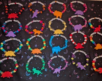 Könnte beinhalten: Eine Sammlung farbenfroher Perlenarmbänder mit dem Text "LOST LANDS" in weißen Buchstabenperlen. Jedes Armband hat einen Dinosaurier-Anhänger in verschiedenen Farben, darunter Rot, Orange und Lila.