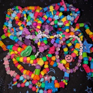 May include: A pile of colorful beaded bracelets with various charms, including smiley faces, stars, hearts, and letters. The bracelets are made with small plastic beads in shades of pink, purple, blue, green, yellow, and orange.