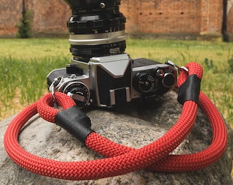 May include: A red camera strap with black accents is wrapped around a silver and black camera with a large lens. The camera is sitting on a grey rock in front of a brick building.