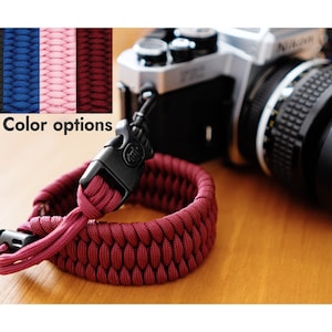 Può includere: Un primo piano di un cinturino per fotocamera in paracord rosso con una fibbia nera. Il cinturino è appoggiato su una superficie di legno accanto a un obiettivo per fotocamera argentato. L'immagine mostra quattro opzioni di colore per il cinturino: nero, blu, rosa e rosso.