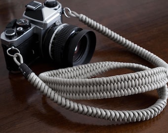 May include: A gray braided camera strap with a silver camera attached. The camera has a black lens with a lens hood.