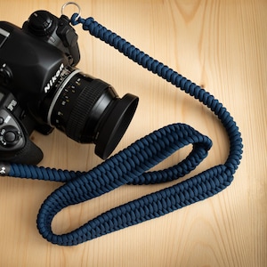May include: A black DSLR camera with a lens attached, sitting on a wooden surface. The camera is connected to a dark blue braided camera strap with silver metal rings.